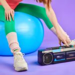 Cropped photo of a woman's legs in tights and leg warmers on an exercise ball, leaning over to turn on a stereo.