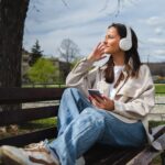 a woman sitting on a park bench wearing headphones and holding a phone in her hand.